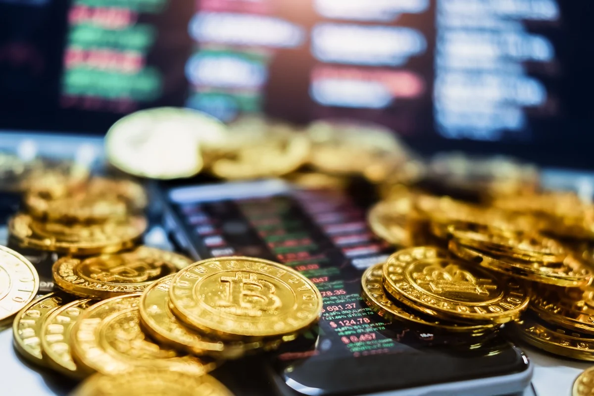 Gold bitcoin coins in front of trading screens, illustrating the SCMP commentary by AADE President Tan Poh Hwee on violent cryptocurrency robberies